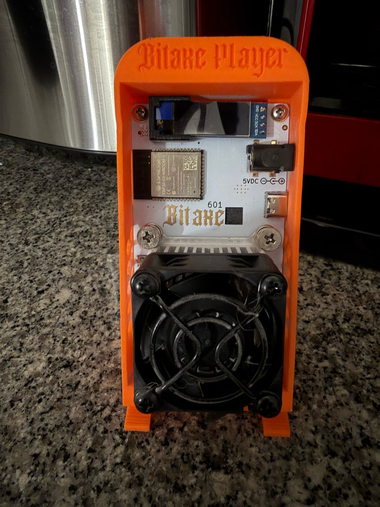 Bitaxe Gamma (Bitcoin Merch), miner solo com alimentação — vista do ASIC no suporte Bitaxe Player